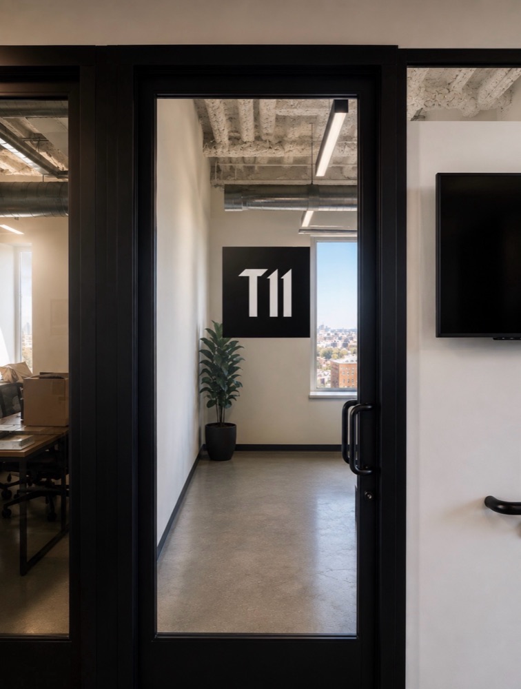 The studio entrance seen through a black-framed glass door, with the T11 mark mounted on the far wall.