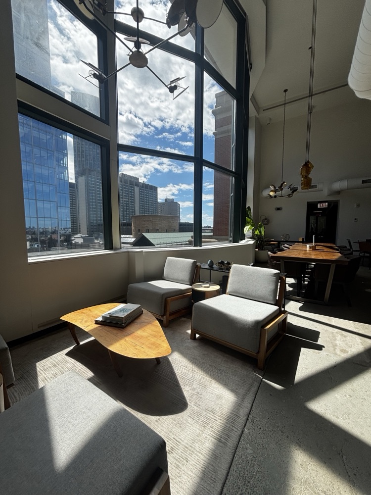 A lounge with low armchairs and a wooden coffee table beside double-height windows looking out over the city.